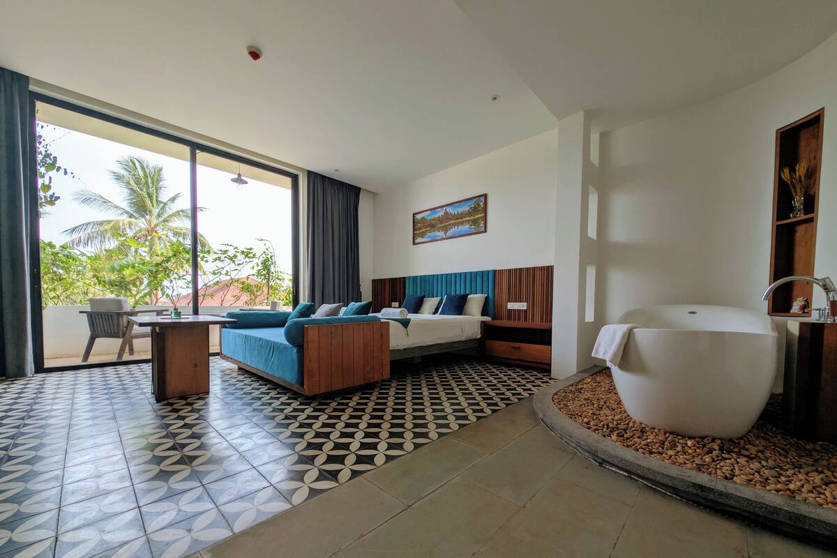 A modern hotel room with a large bed, a sofa, a dining table, and a bathtub.