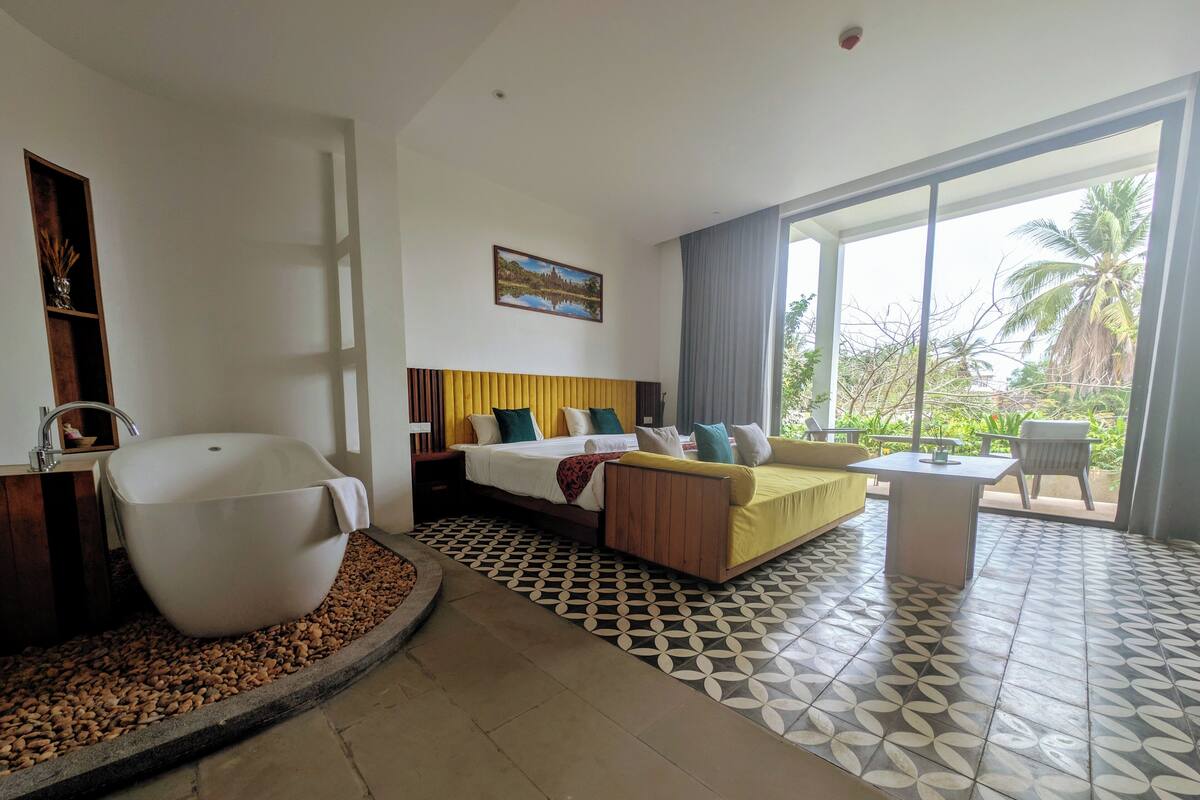 A hotel room with a bed, a large white bathtub, a dining area, and a view of palm trees.