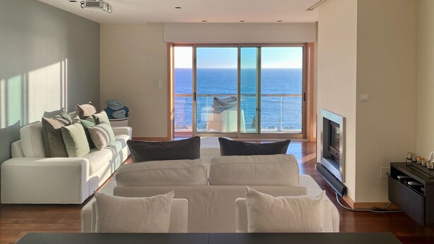 A modern living room with a large window offering a view of the ocean. There is a white sofa with multiple cushions, a flat-screen TV, and a small table. - Luxury Apartment on Lavadores Beach (Vila Nova de Gaia)