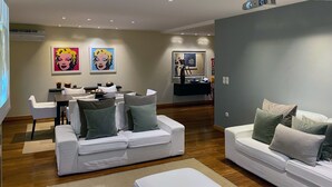 A modern living room with two white sofas, a coffee table, and a dining area with a table and chairs. There are two framed artworks on the wall, and a projector screen is visible. - Luxury Apartment on Lavadores Beach (Vila Nova de Gaia)