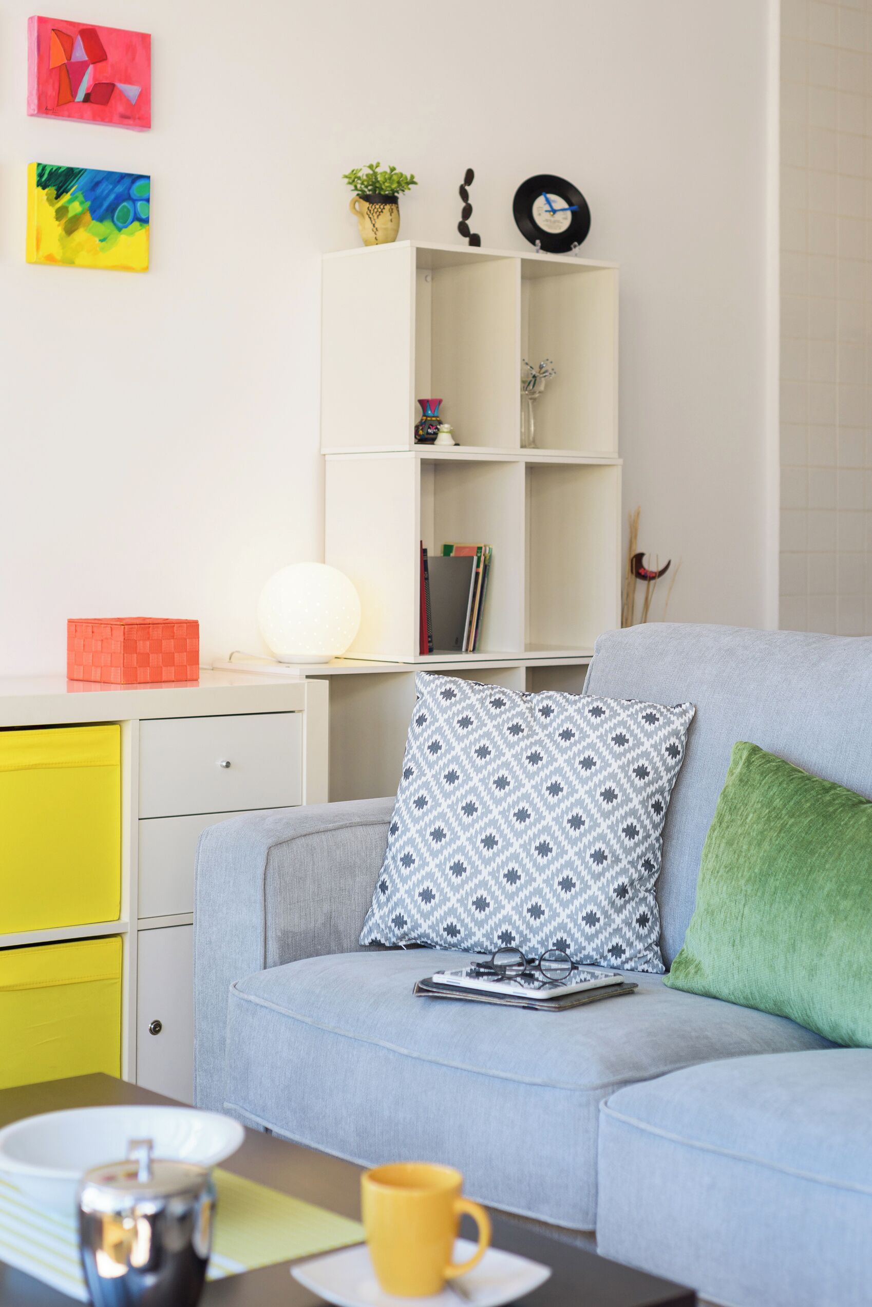 Uma sala de estar moderna com um sofá cinza, um armário amarelo, uma estante branca com livros e objetos decorativos, e uma pintura abstrata vermelha e azul na parede.