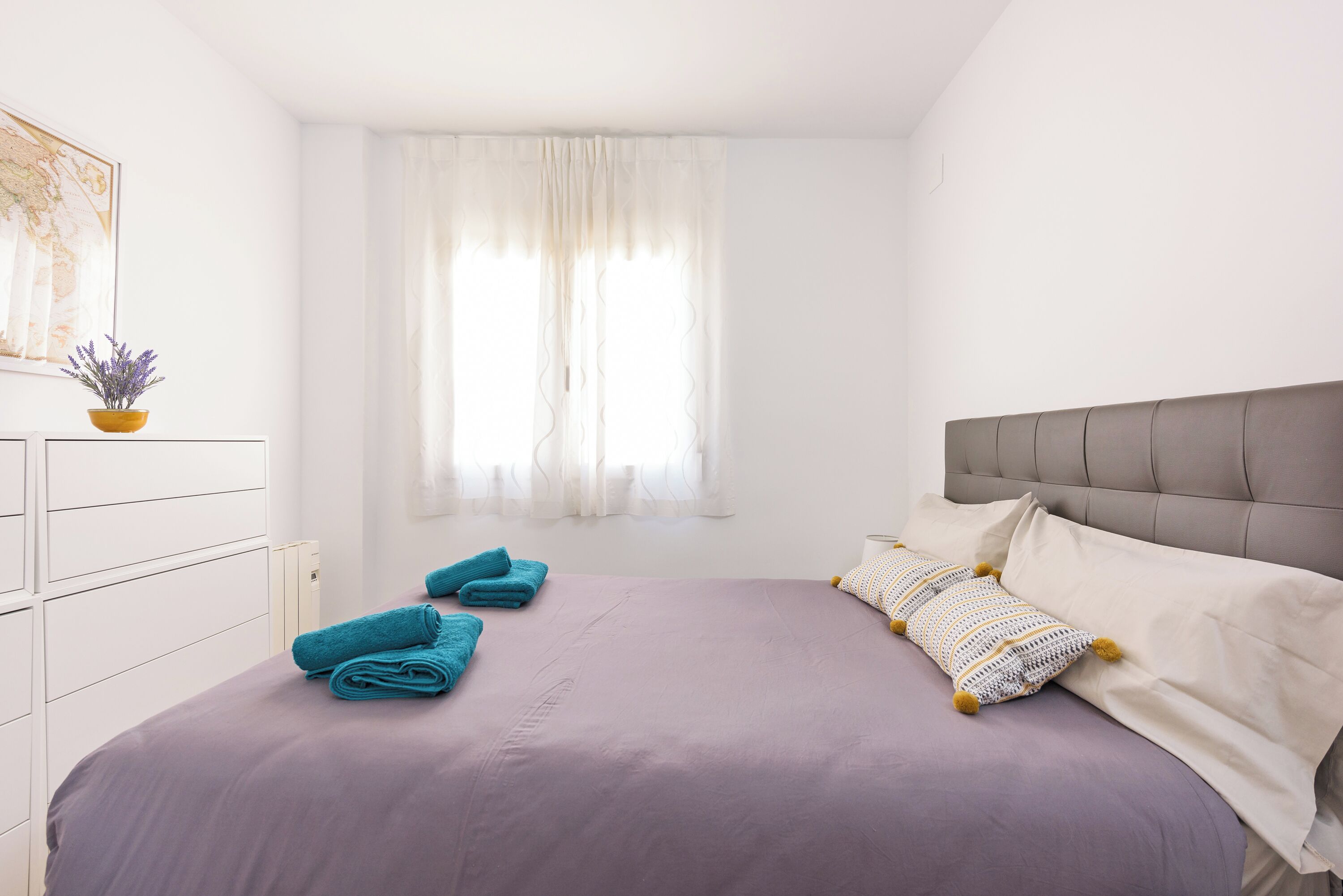 A bedroom with a bed, pillows, a nightstand, a window with curtains, a radiator, and a framed picture on the wall.