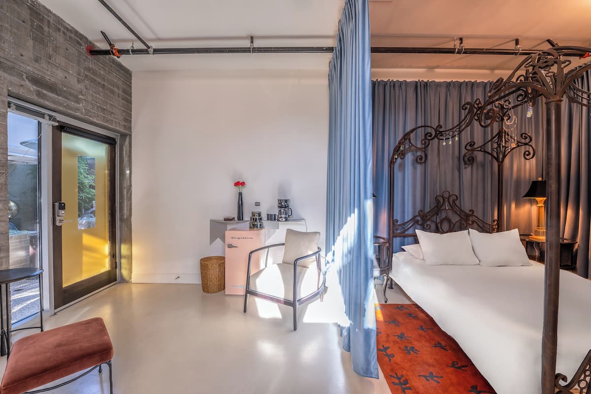 A modern hotel room with a large bed, a desk, and a chair. There is a window with curtains, a small table with a vase of flowers, and a red rug.