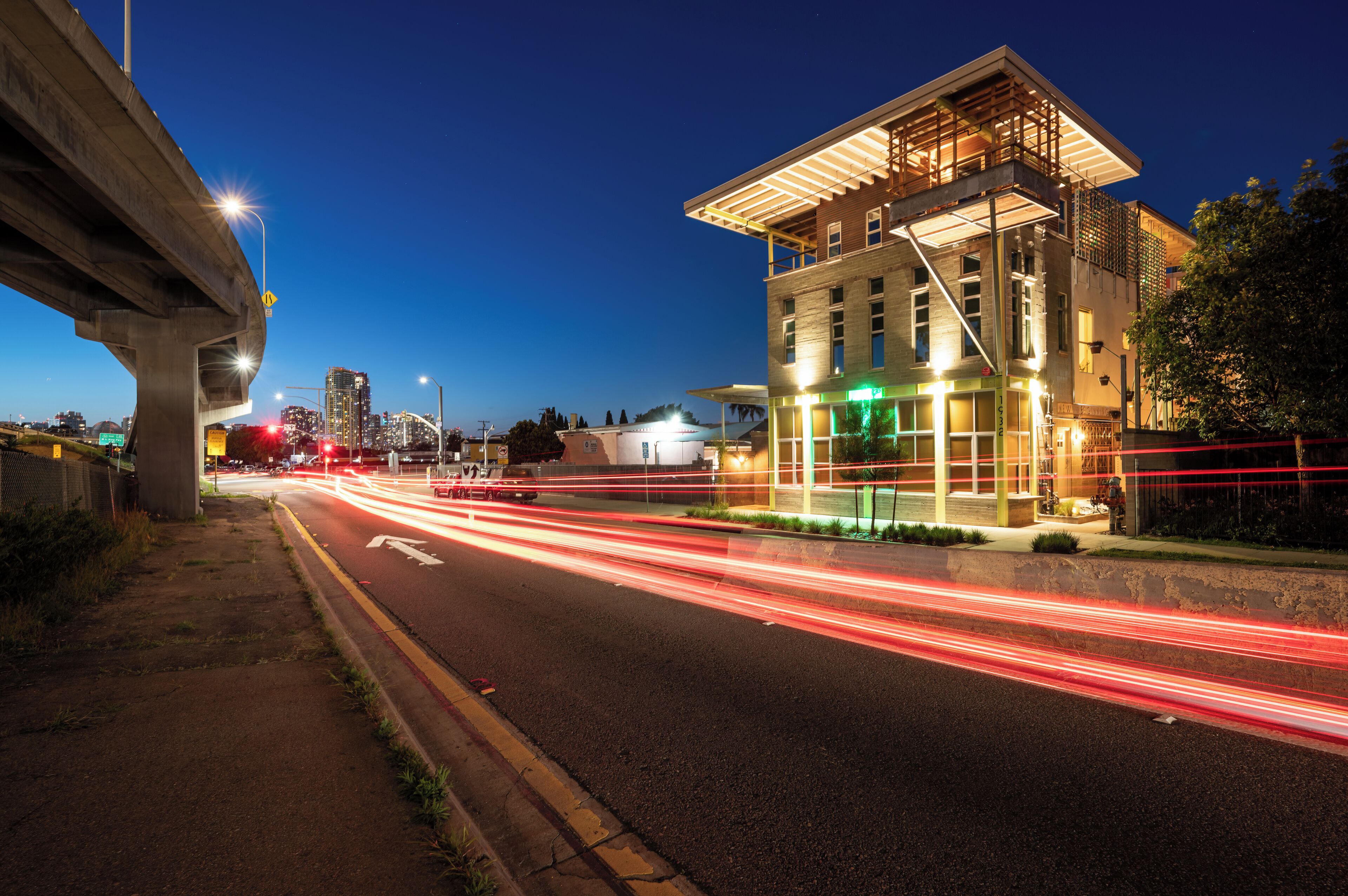 A modern building with a flat roof and large windows, situated next to a busy road with light trails from moving vehicles.