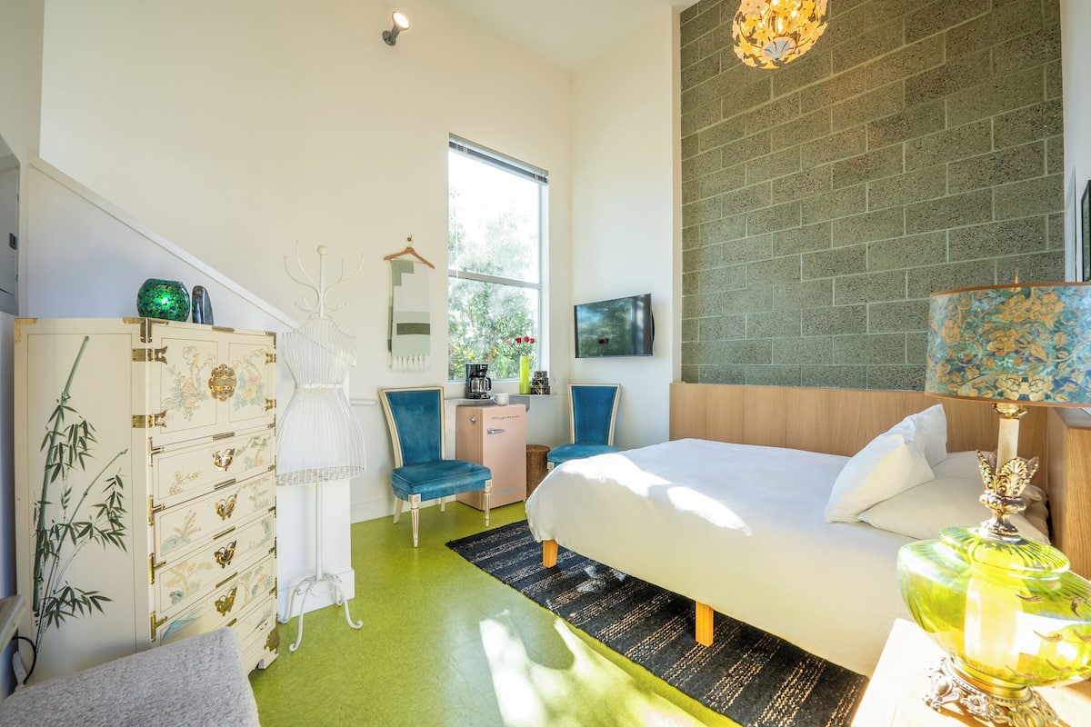 A modern bedroom with a large bed, a dresser with decorative elements, a desk with a chair, and a large window providing natural light.