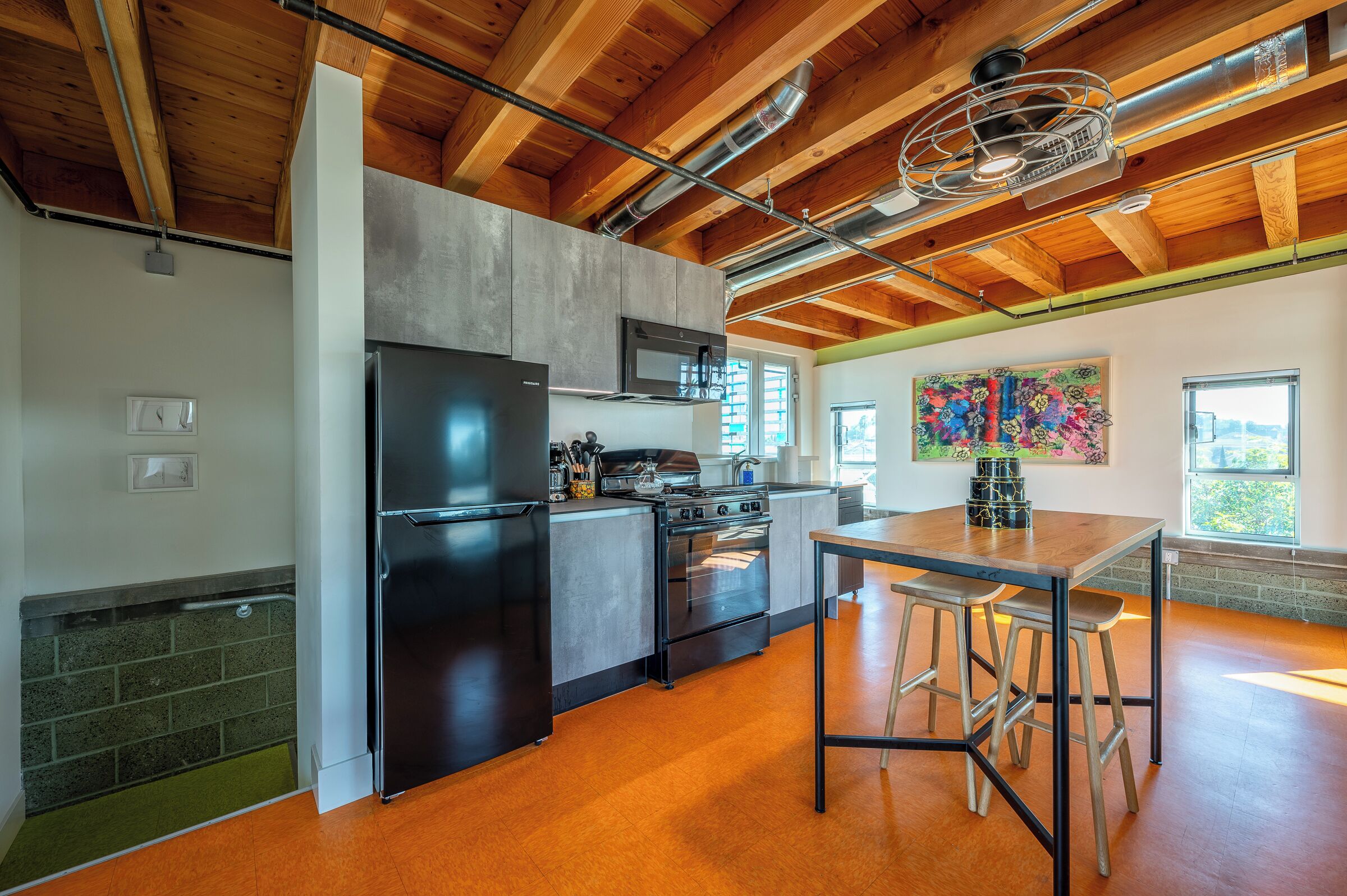 A modern kitchen with a stainless steel refrigerator, oven, and microwave, wooden ceiling beams, and a dining area with stools.