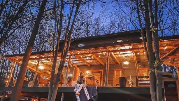 Une cabane en bois avec un porche couvert, une personne assise sur le porche et une vue sur les arbres environnants.
