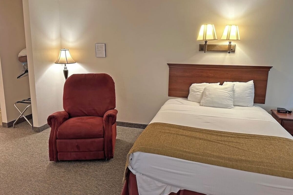 Una habitación de hotel con una cama, un sillón rojo, dos lámparas de pared y una mesita con lámpara.