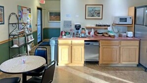 A small kitchen with wooden cabinets, a microwave, a coffee maker, and a sink. There is a small table with chairs and a bulletin board with papers. - Vision Inn Motel (Falls City)