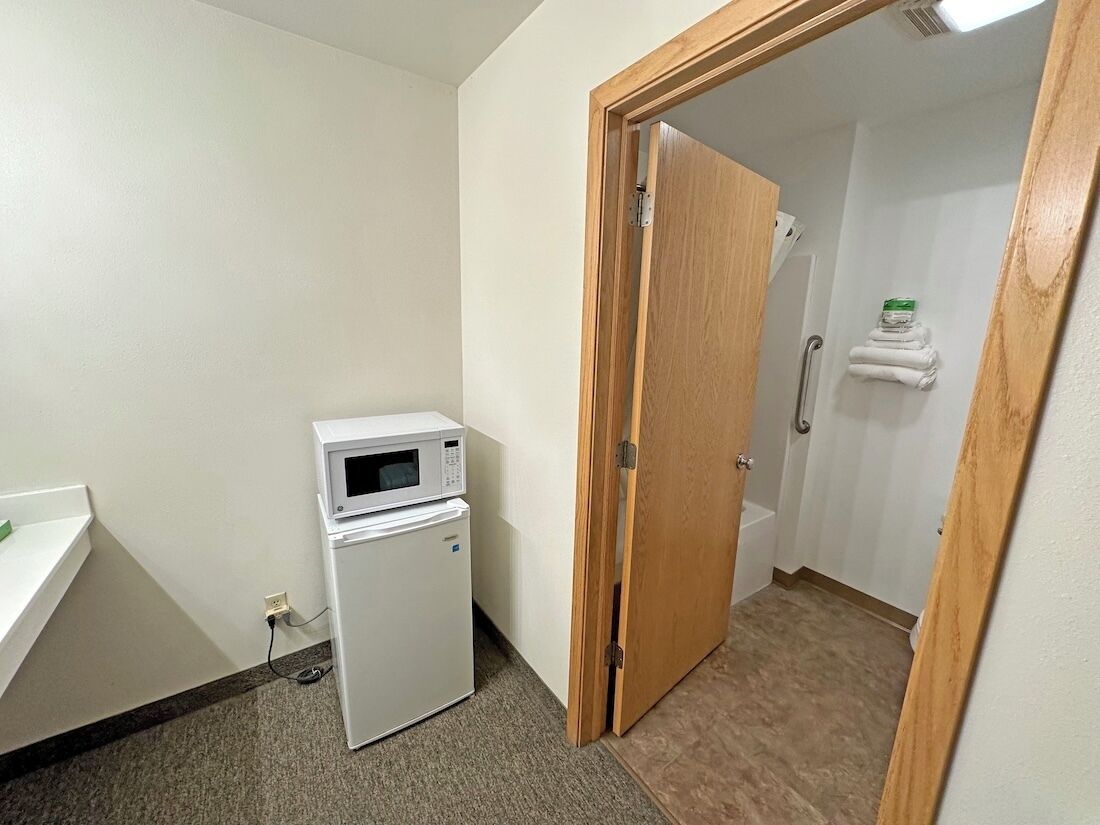 Una habitación pequeña con microondas, refrigerador, una puerta de madera y un perchero con toallas.
