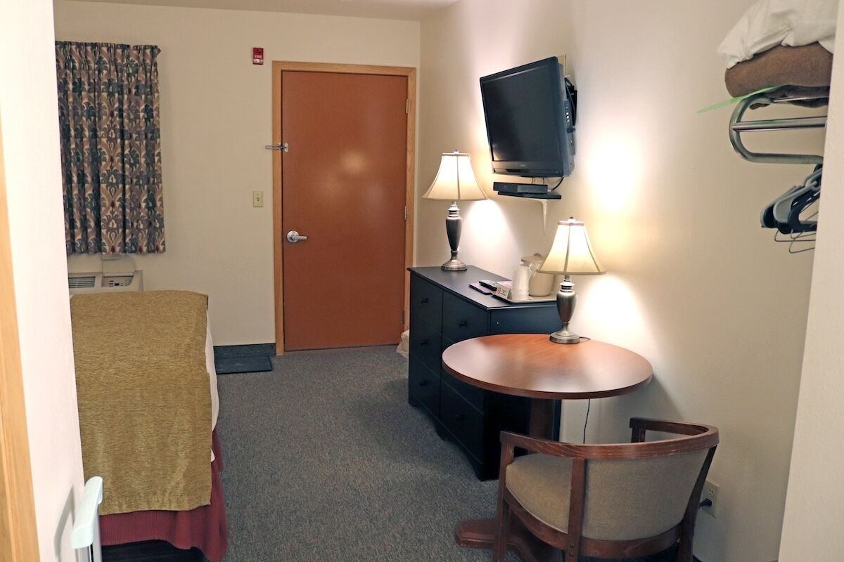 A compact hotel room with a bed, a small table with two lamps, a television on a stand, and a coat rack with hanging items.