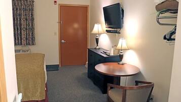 A compact hotel room with a bed, a small table with two lamps, a television on a stand, and a coat rack with hanging items.