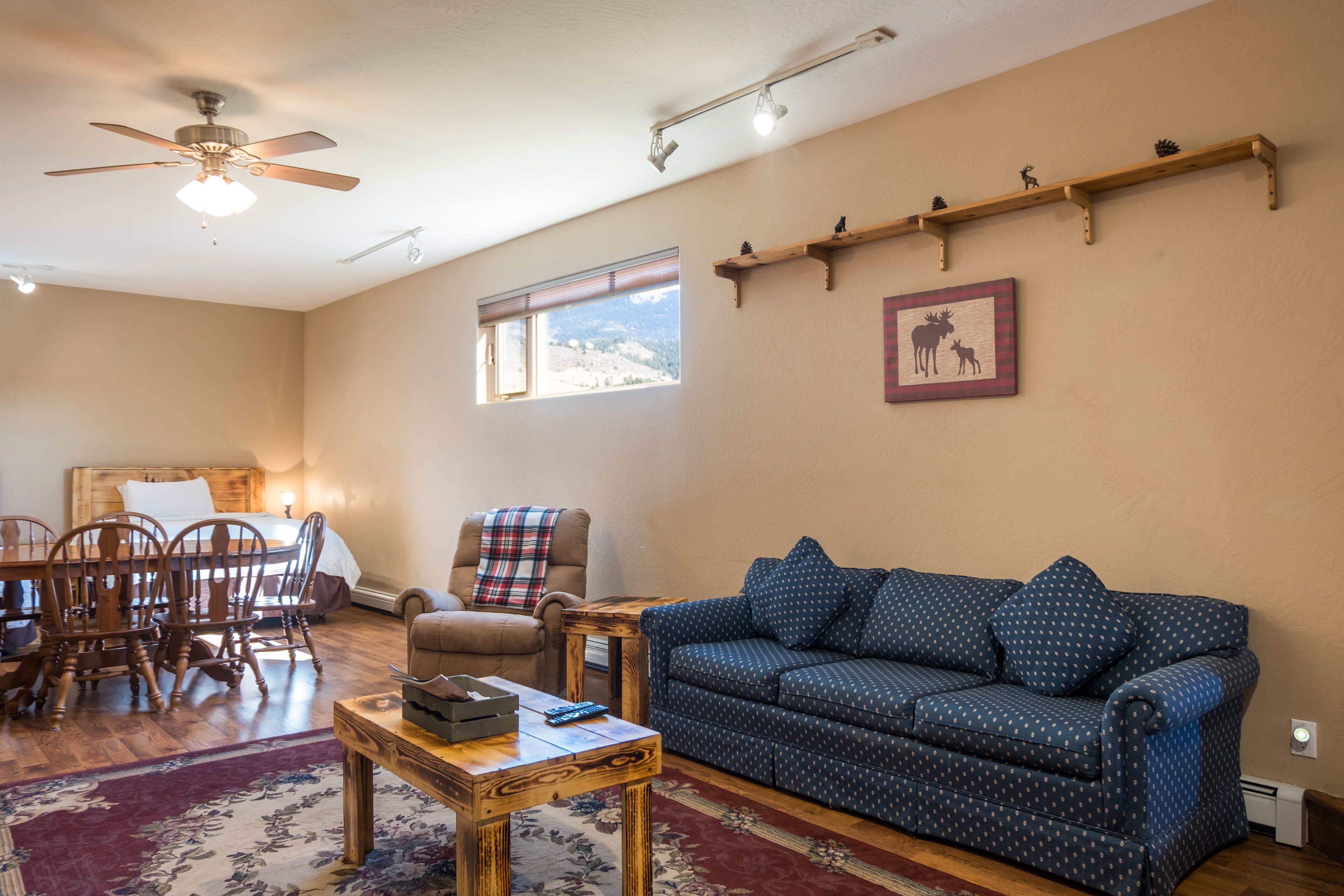 A cozy living room with a blue patterned sofa, wooden chairs, a wooden coffee table, a plaid armchair, and a ceiling fan.