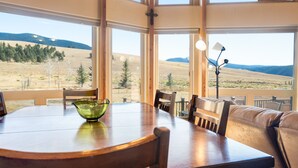 A dining area with a wooden table and chairs, a green glass bowl, and a view of a mountainous landscape through large windows.