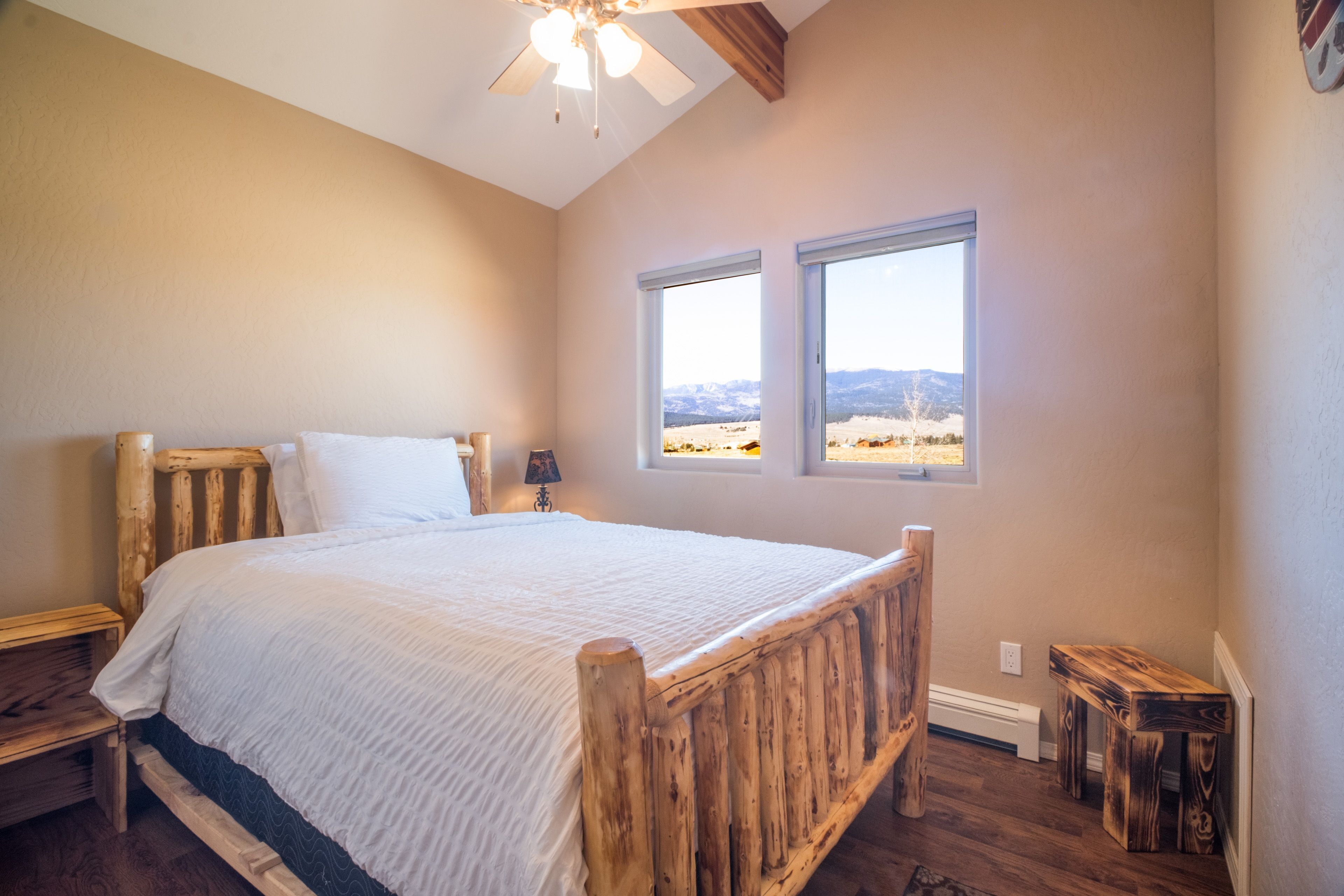 A bedroom with a wooden bed, two windows with a mountain view, a ceiling fan, and a small bedside table.
