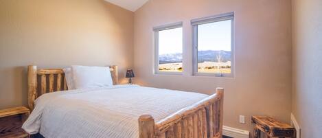A bedroom with a wooden bed, two windows with a mountain view, a ceiling fan, and a small bedside table.