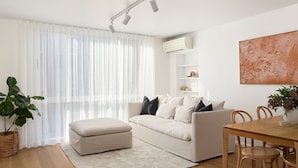 Un salon moderne avec un canapé blanc, une table à manger avec des chaises, une grande plante en pot et un tableau accroché au mur.