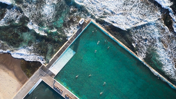 Vue aérienne d’une piscine rectangulaire à l’eau d’un bleu limpide, bordée d’une plage de sable et d’une côte rocheuse.