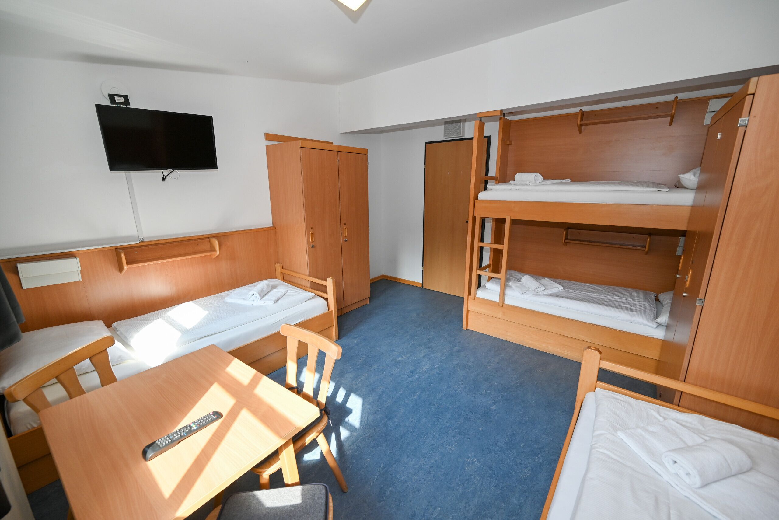A room with bunk beds, a TV mounted on the wall, a wooden wardrobe, and a table with a remote control.