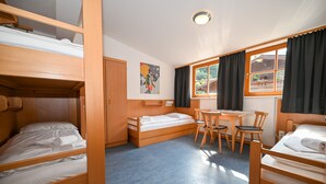 Ein Zimmer mit Etagenbetten, einem kleinen Tisch, Stühlen und einem Fenster mit Vorhängen.