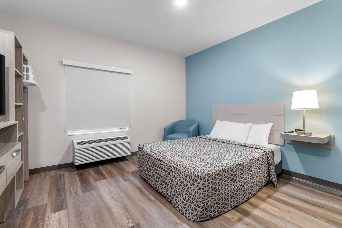 Una habitación de hotel moderna con cama, una silla, mesita de noche con lámpara, armario y aire acondicionado instalado en la pared.