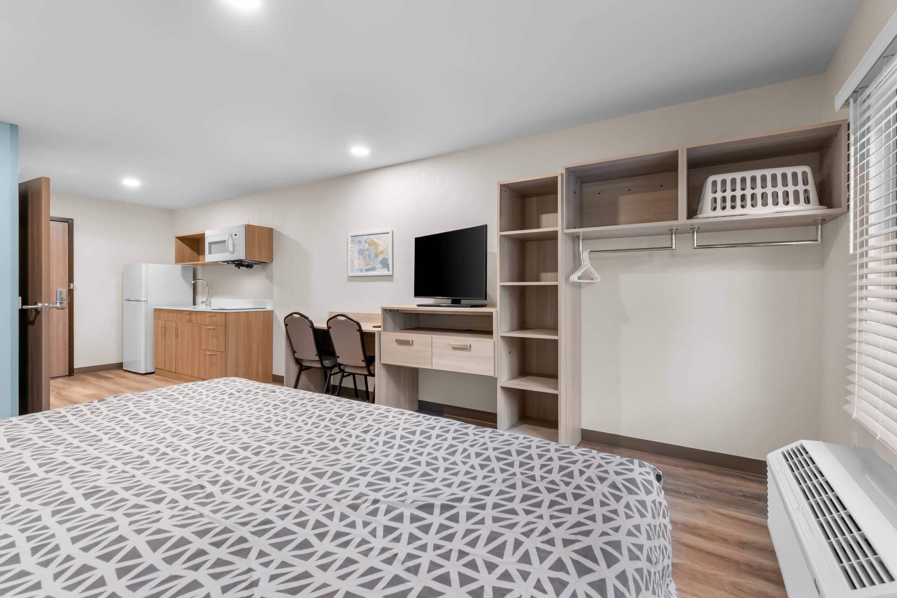 A bedroom with a bed, a desk, a TV, and a kitchenette.