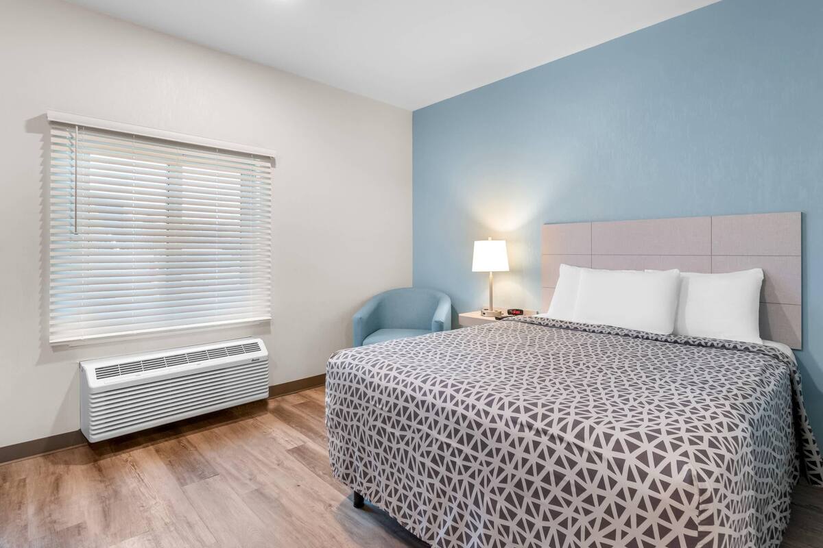Habitación de hotel con cama, una silla, mesita de noche, lámpara y ventana con persianas.