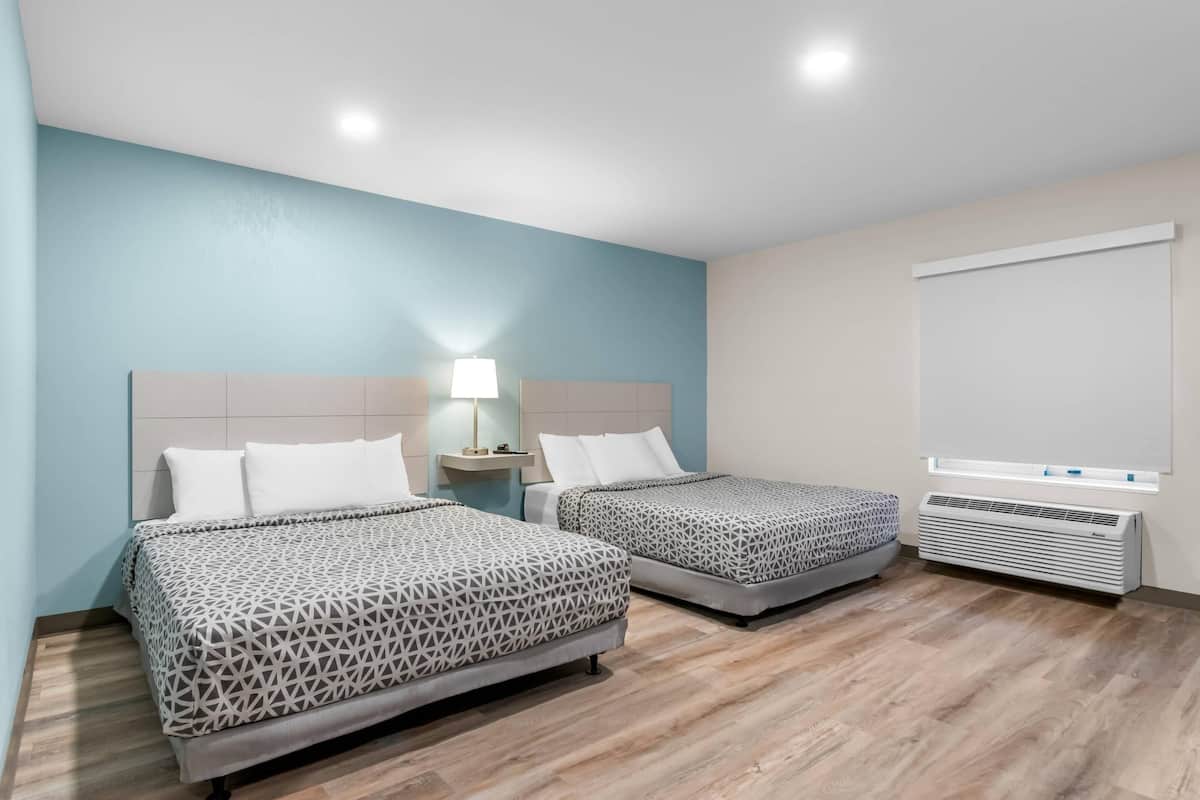 Habitación de hotel con dos camas, una mesita de noche con lámpara, una ventana con persianas y un aire acondicionado instalado en la pared.