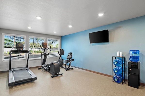Un gimnasio con cintas de correr, elípticas y bicicletas estáticas. Hay ventanas amplias con persianas, un dispensador de agua y un televisor montado en la pared.