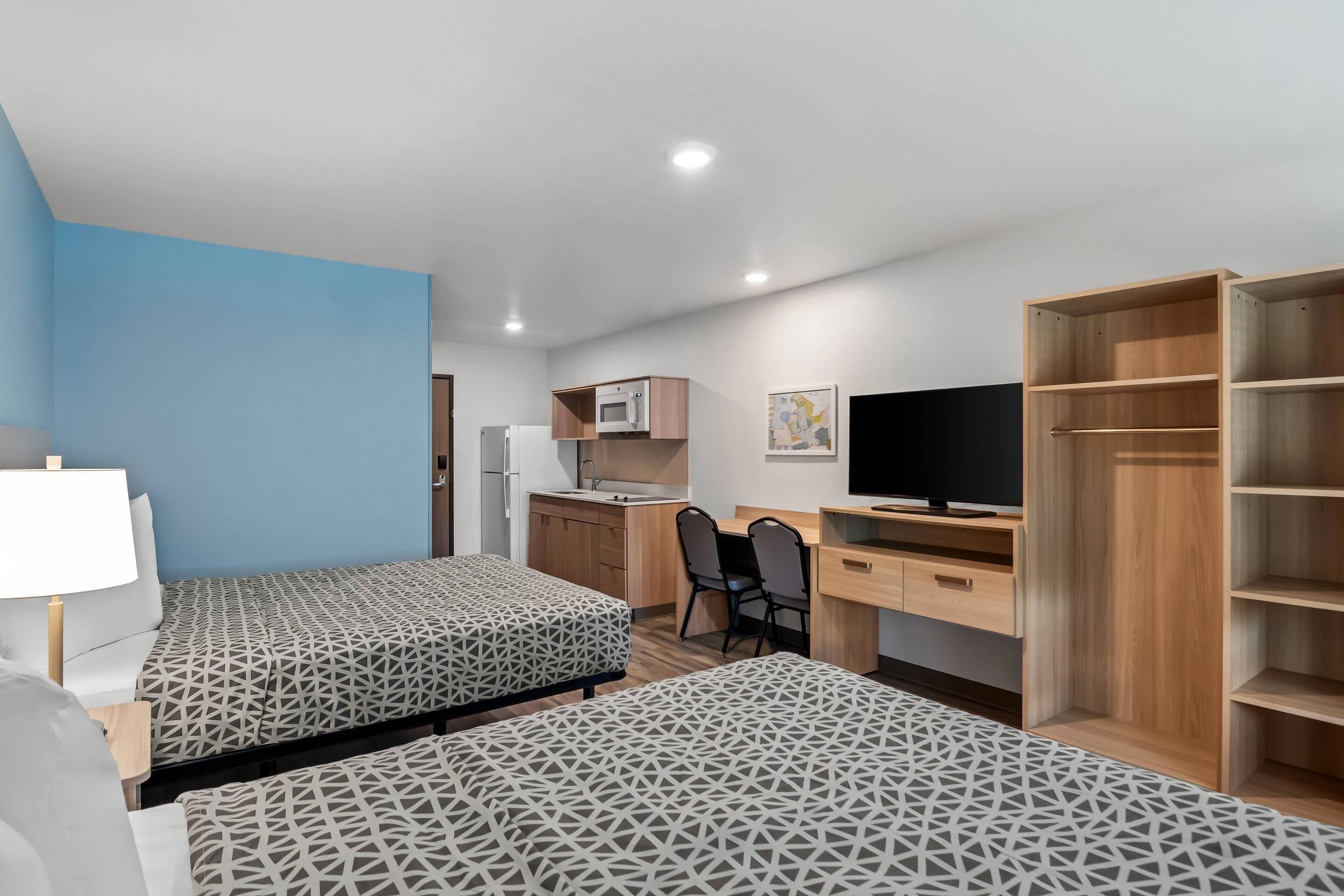 A compact hotel room with two beds, a desk, a TV, a kitchenette, and a wardrobe.