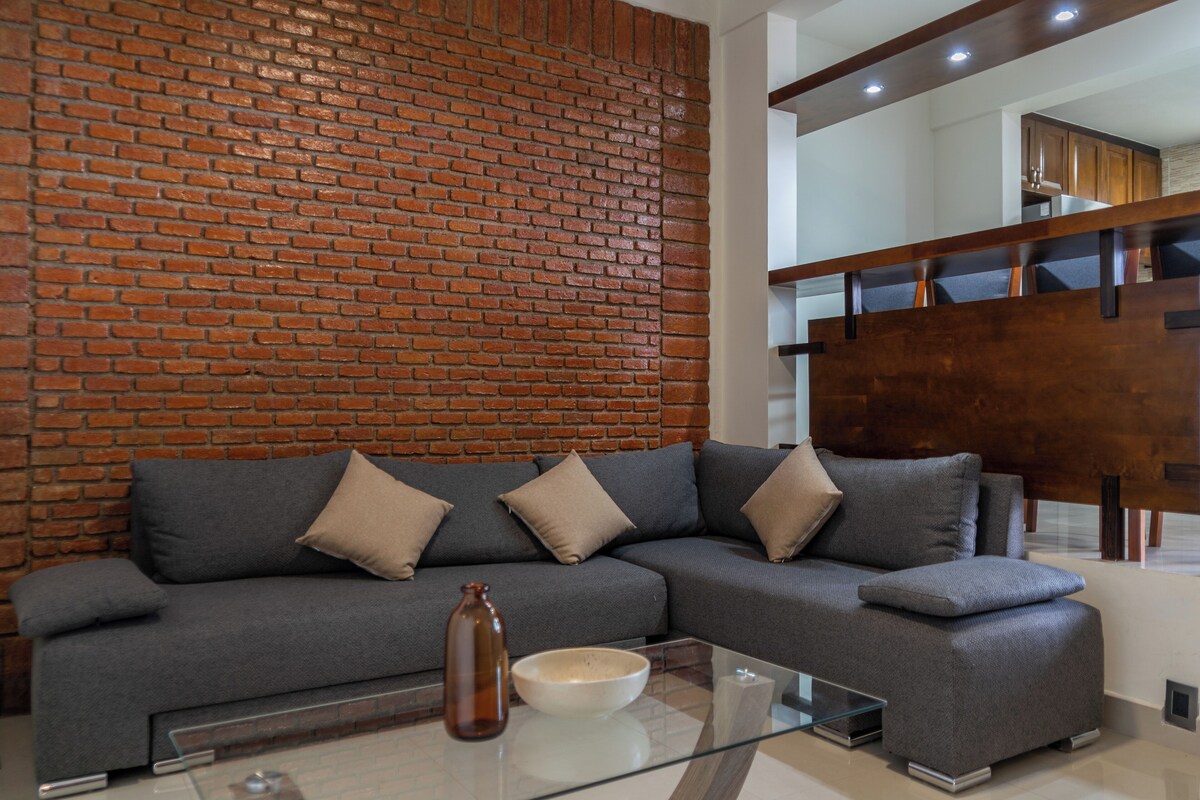 A modern living room with a grey sectional sofa, a glass coffee table, and a brick accent wall.