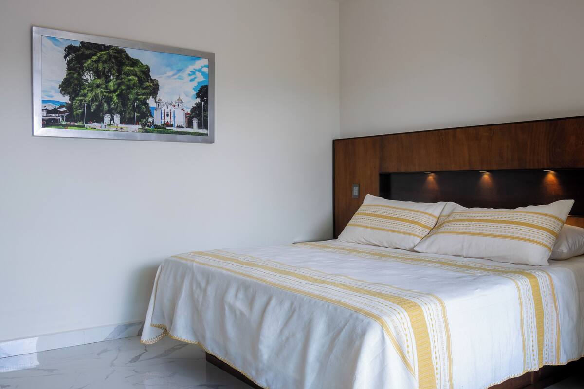 Ein ordentlich bezogenes Bett mit einer weiß-gelb gestreiften Tagesdecke, einem Holzkopfschild und einem gerahmten Bild eines Gebäudes an der Wand.