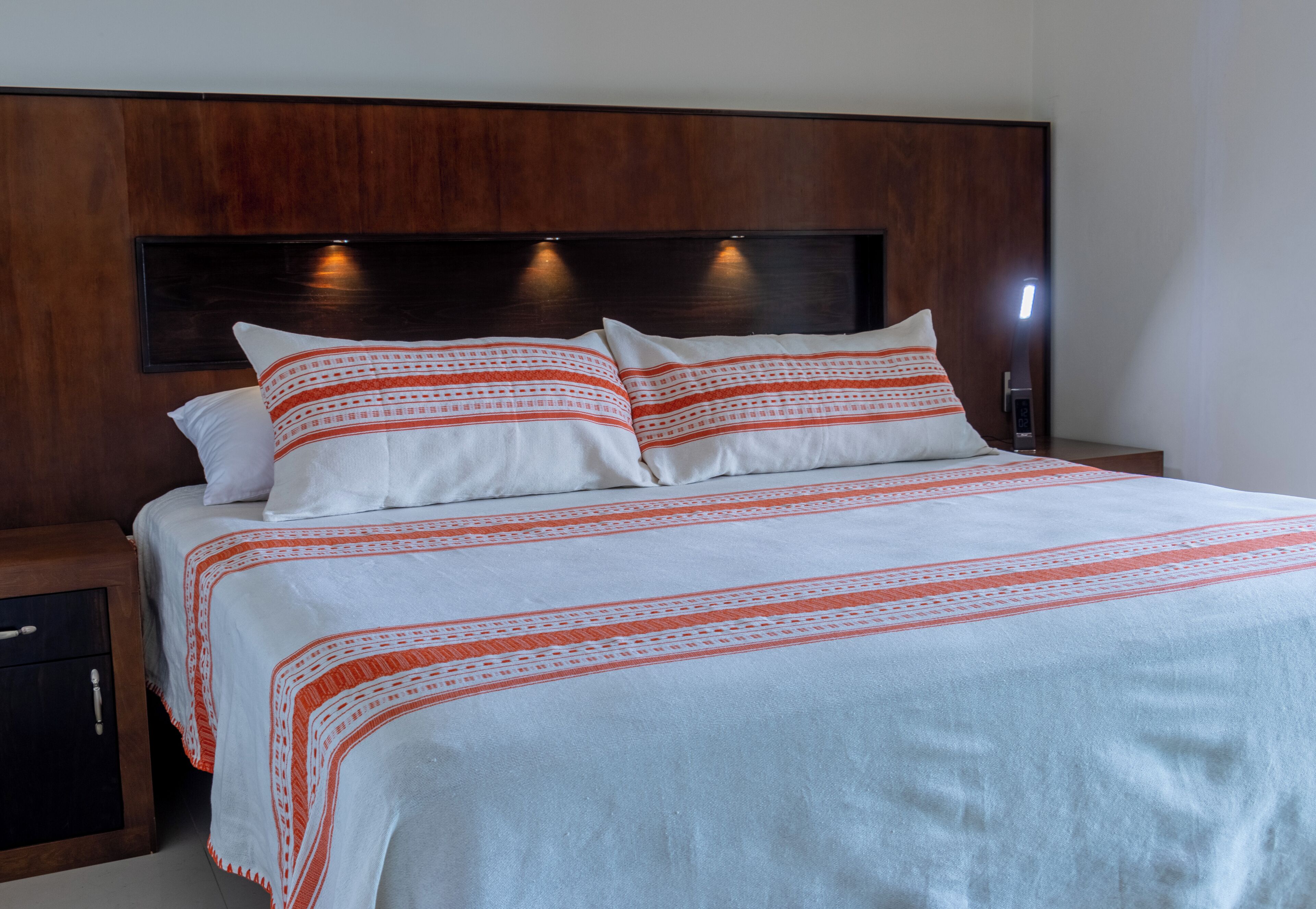 A bed with white bedding and red patterns, a wooden headboard, and a nightstand with a drawer.