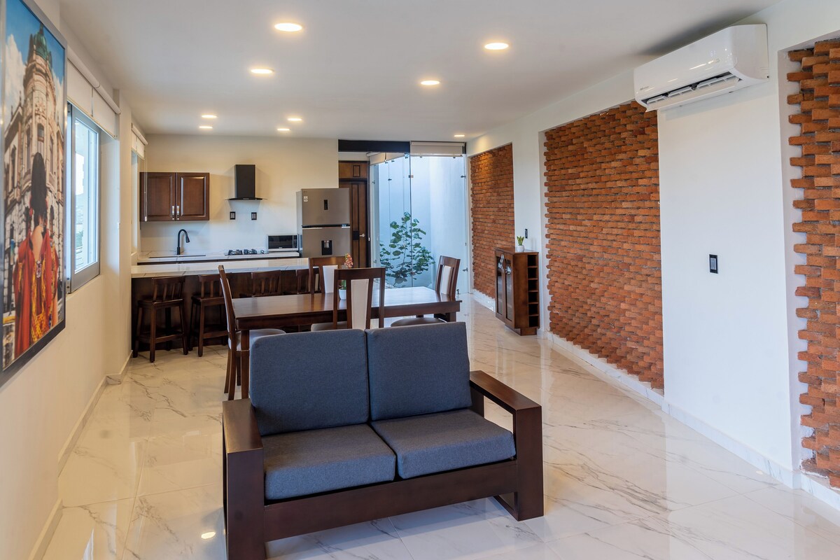 A modern living room with a sofa, dining area, kitchen, and a brick wall.