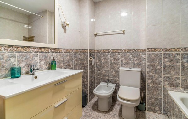 A bathroom with a marble-tiled wall, a sink with a mirror, a bidet, and a toilet.