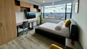 A modern bedroom with a bed, a desk, a TV, and a large window with a city view.