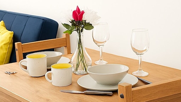 A wooden dining table set with white ceramic dishes, two white mugs, a vase with flowers, and two wine glasses.