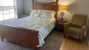 A bedroom with a wooden bed, a nightstand with a lamp, a chair, and a window with blinds.