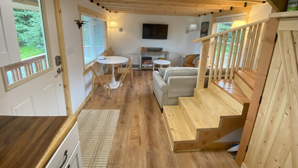 A cozy interior with a wooden staircase, a dining area with a round table and chairs, a sofa, and a TV mounted on the wall. - Rustic Homer Cabin w/ Moose Views & Quiet Porch (Homer)