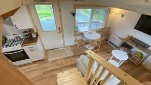 A compact kitchen with a gas stove, oven, and white cabinets. There is a small dining area with a table and chairs, and a balcony with a railing. - Rustic Homer Cabin w/ Moose Views & Quiet Porch (Homer)