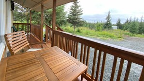 A wooden deck with a table and chairs, overlooking a forested area with a gravel path and a body of water in the distance. - Rustic Homer Cabin w/ Moose Views & Quiet Porch (Homer)
