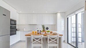 Une cuisine moderne avec des meubles blancs, un plan de travail en bois et deux chaises blanches. On y trouve un réfrigérateur, un four et un micro-ondes sur le côté gauche. Une plante et un saladier de fruits sont posés sur le plan de travail.
