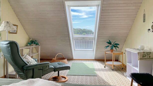 A cozy attic room with a sloped ceiling, a green cushioned chair, a small round table with a potted plant, and a view through a large window.