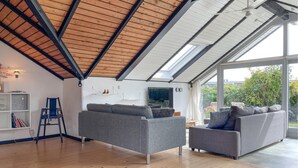 A spacious room with a high ceiling, wooden beams, a grey sofa, a smaller grey armchair, and a large window offering a view of greenery. - Awesome home in Tranekær with WiFi (Tranekær)