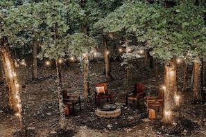 Un décor forestier avec des chaises en bois disposées autour d’un foyer central, le tout entouré d’arbres et illuminé de guirlandes lumineuses.