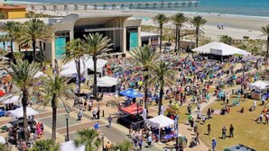 Un evento al aire libre concurrido con un escenario y carpas, palmeras y una playa al fondo.