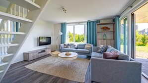 A modern living room with a grey sofa, a coffee table, and a TV on a white cabinet. There is a staircase on the left and a large window with curtains. - 3 bedroom amazing home in Kamperland (Kamperland)