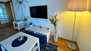 A modern living room with a flat-screen TV mounted on the wall, a round clock, a coffee table, a TV stand, a lamp, and a vase with flowers. - Tejo Balcony Expo (Lisbon)