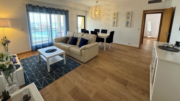 A modern living room with a sofa, dining table, and chairs, a chandelier, and a large window with curtains. - Tejo Balcony Expo (Lisbon)