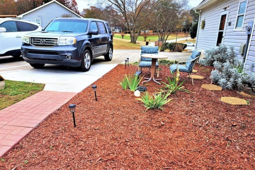 A residential driveway with a parked SUV, a patio area with outdoor furniture, and a landscaped garden bed.